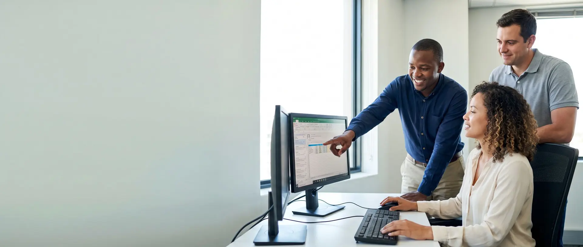 A smiling IT technician points at a colleague’s monitor while explaining a fix, as another colleague looks on in a bright office